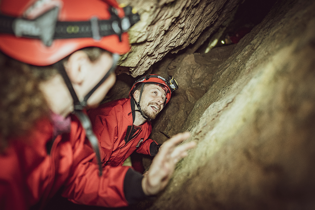 A man in a  red boiler suit and helmet looks upwards as he squeezes through a small crevice in a cave